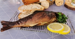 Eine goldbraune, geräucherte Forelle liegt mit 2 Stücken Brot, 2 Zitronenscheiben und etwas Petersilie dekoriert auf einem Glasteller.
