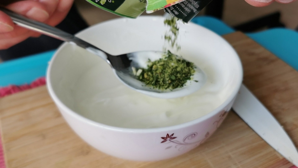 Ein Knoblauchdip wird in einer Schüssel aus griechischem Joghurt und Quark angerührt und mit einem Löffel grüner Gartenkräuter veredelt