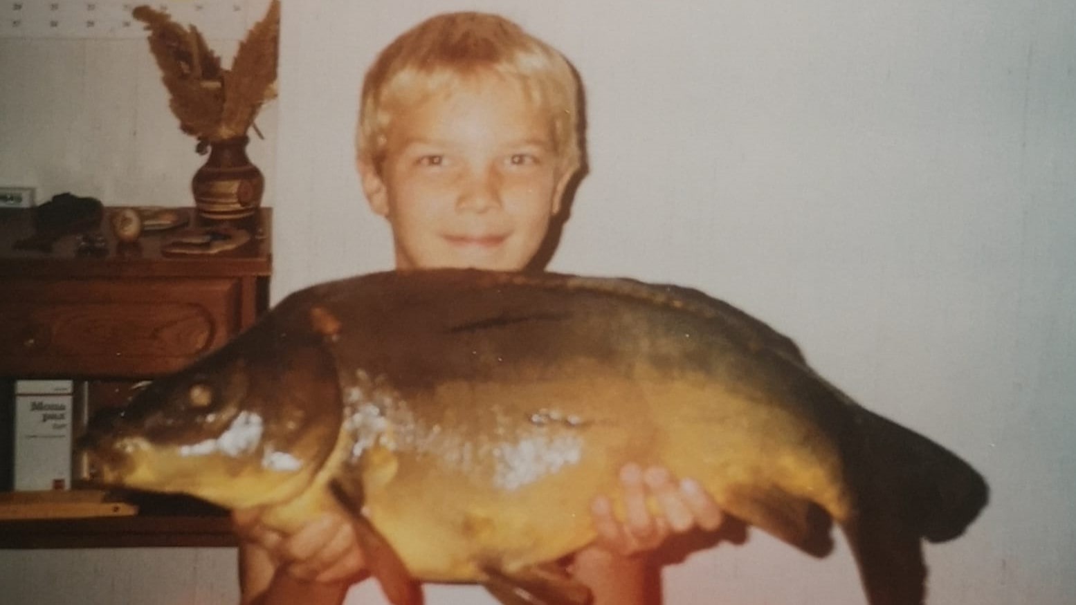 In den 80er Jahren hält ein kleiner blondhaariger Junge freundlich lächelnd einen stattlichen Karpfen in den Händen, stolz auf seinen erfolgreichen Fang.