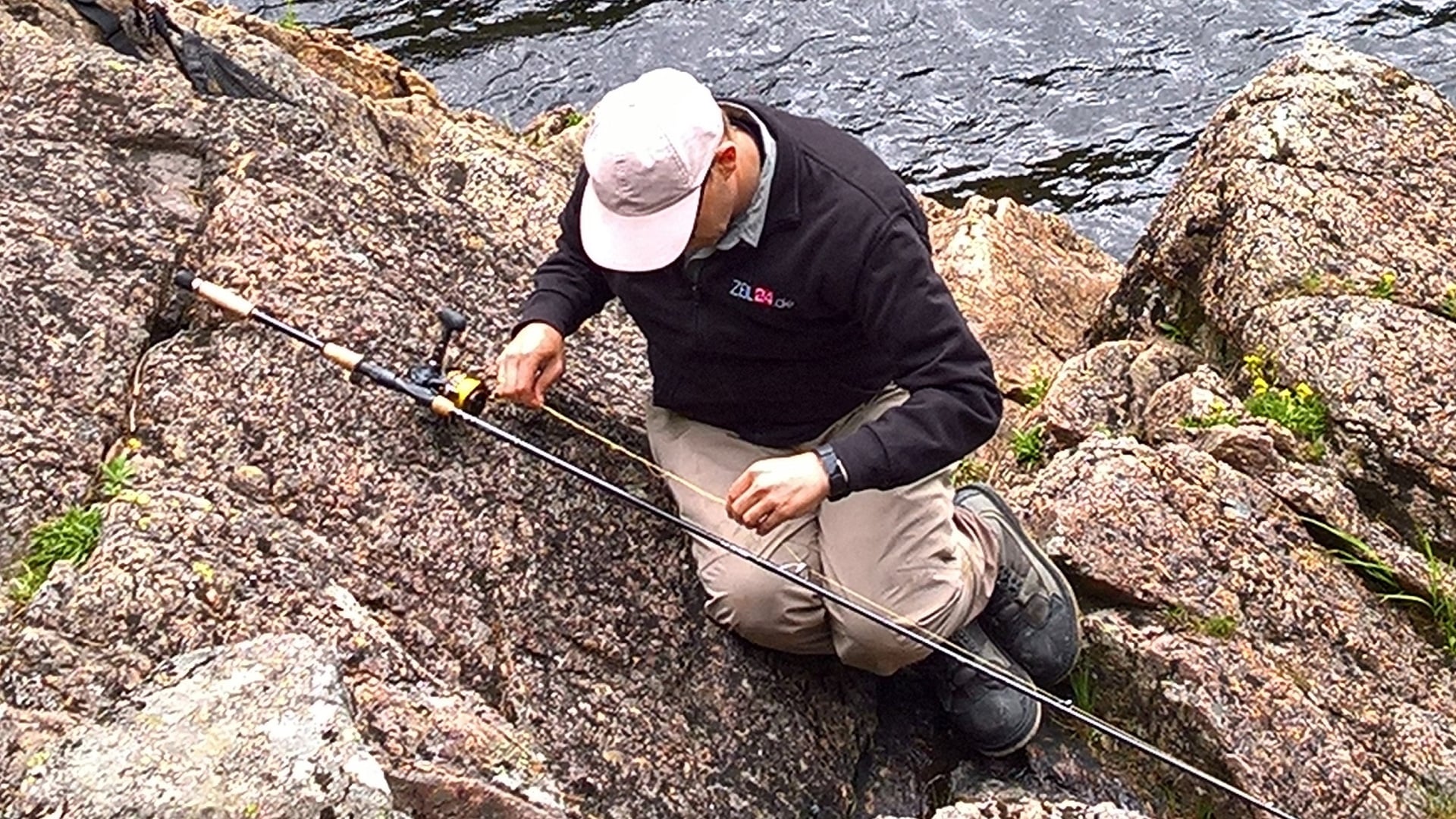 Ein Angler sitzt auf dem felsigen Ufer eines Lachsflusses und untersucht den Zustand des Fluorocarbon-Vorfachs an seiner Angelrute.