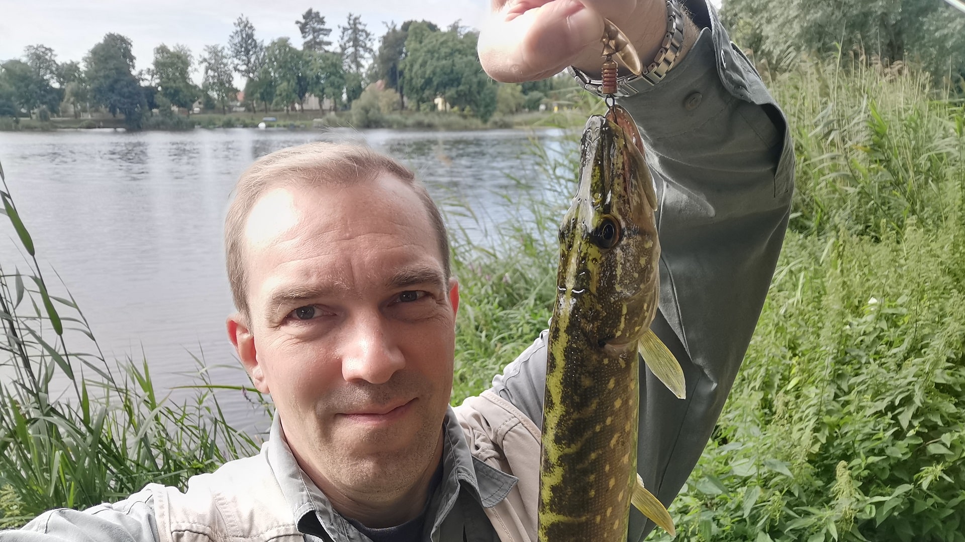 Ein Angler präsentiert stolz einen kleinen Hecht in die Kamera. Hintergrund: grünes Schilf am Nordufer des Mühlensees in Liebenwalde.
