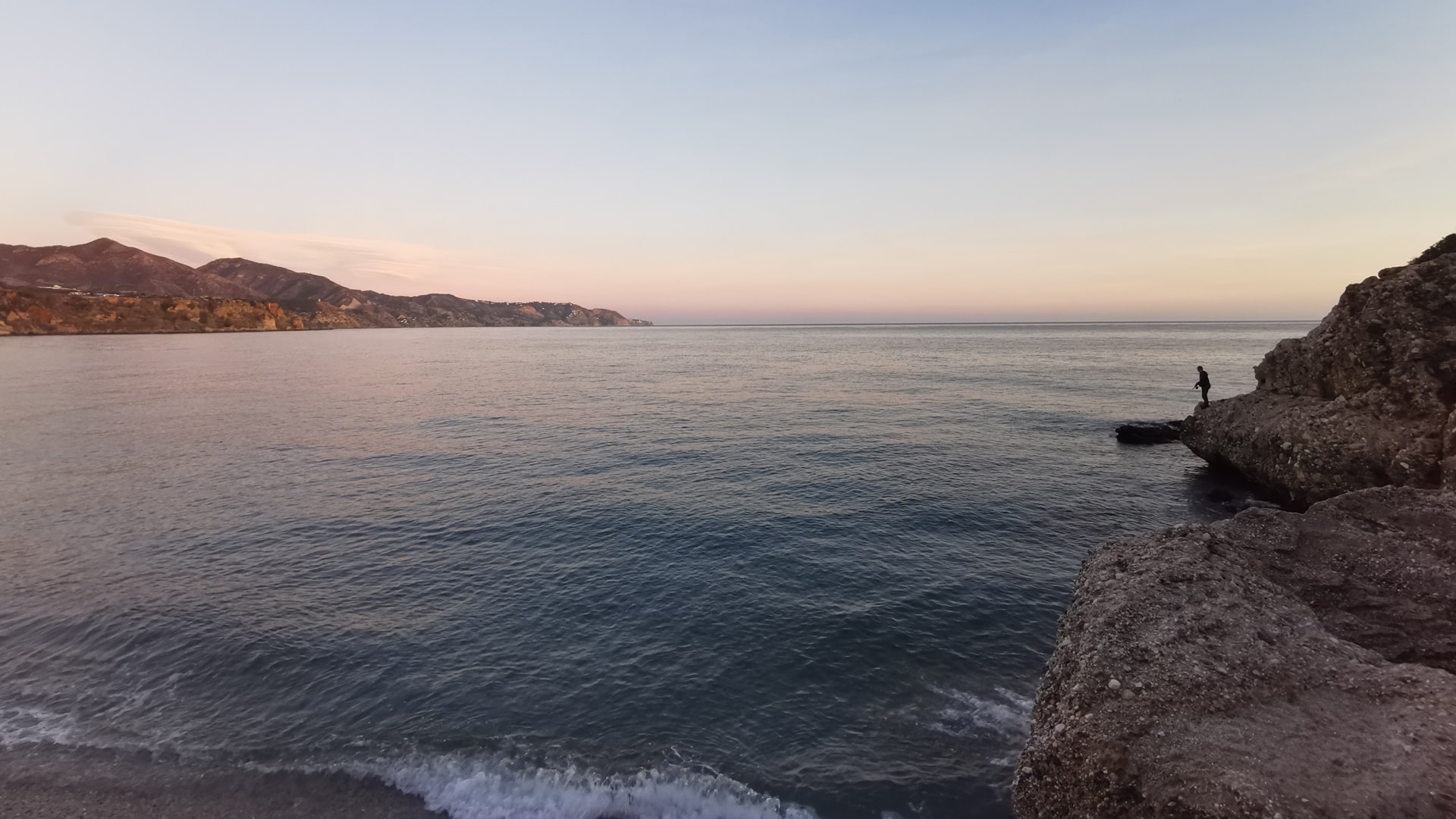 Abendstimmung in Nerja, einer Küstenstadt in Andalusien, Spanien, mit Blick auf das Mittelmeer, die braune Felsenküste, einen Angler, der sein Glück mit der Spinnrute versucht, und einen klaren, hellblauen Himmel.