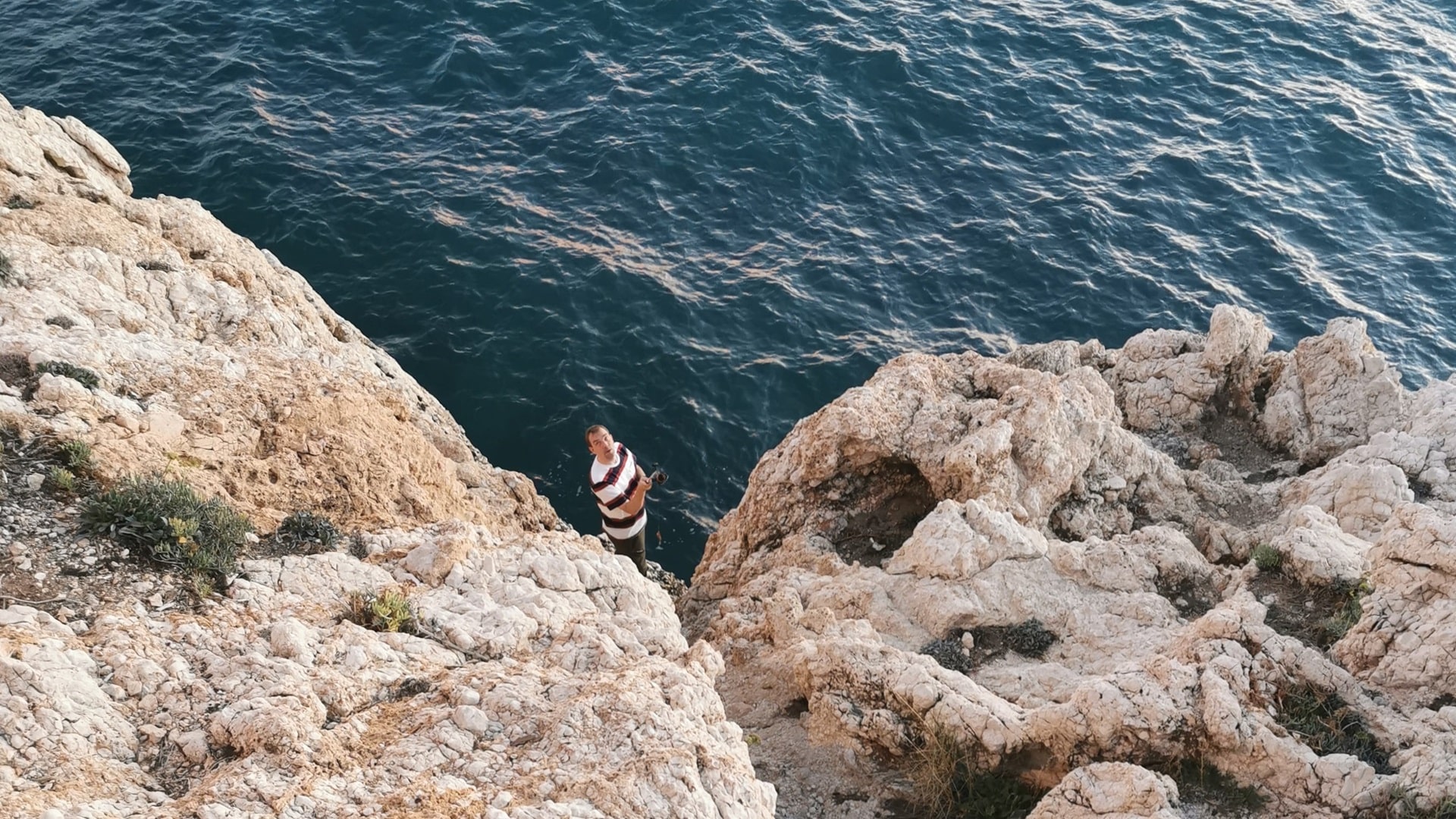 Beim Spinnfischen 5 Meter unter dem Holzgeländer des Uferweges zwischen Rincón de la Victoria und Cala del Moral bei Malaga, Andalusien, steht ein Angler auf weißen Felsen, blickt die steile Wand hinter sich hinauf - vor ihm das ruhige Meer