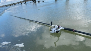 Eine fertig montierte lange Meerforellenrute liegt mit einer salzwassertauglichen Rolle bei kühlen Temperaturen unter blauem Himmel am Strand einer Meerforellenbucht.