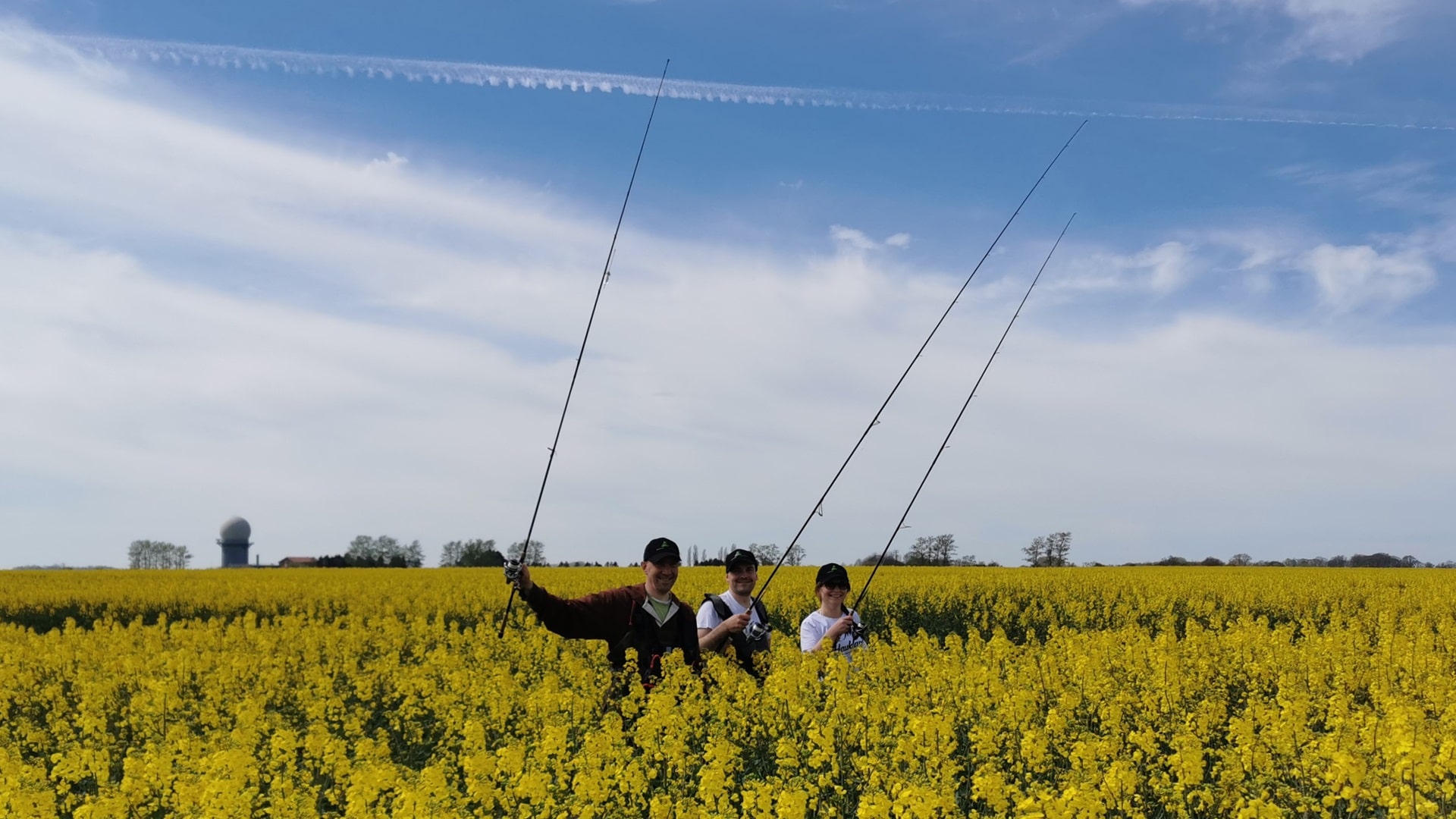 Drei Angler stehen inmitten eines gelb blühenden Rapsfeldes und strecken voller Vorfreude ihre Ruten in die Höhe, da die Zeit, wenn der Raps blüht, signalisiert, dass die Saison zum Hornhechtangeln gekommen ist.