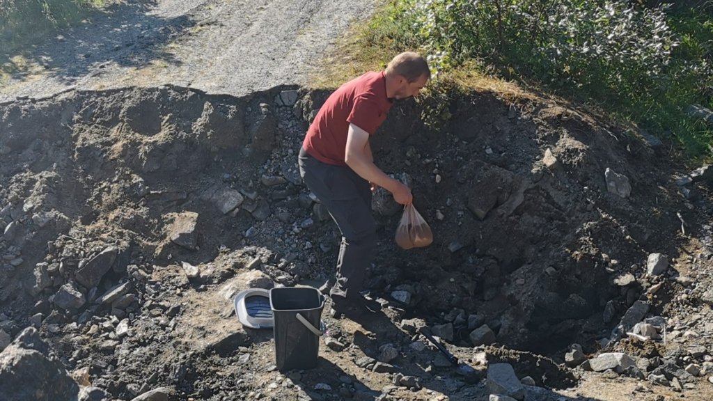 Ein Mann legt einen kompostierbaren Beutel mit Exkrementen in ein 20cm tiefes Loch, dass er vorher mit einem Klappspaten gegraben hat, um es danach zuzuschaufeln