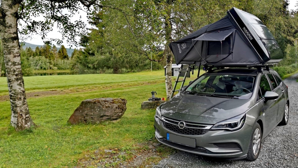 Ein aufgebautes Dachzelt auf einem gepflegten Schotterparkplatz bei Klæbu, daneben eine saftig grüne Wiese, an die ein See anschließt, in dem man Angeln kann