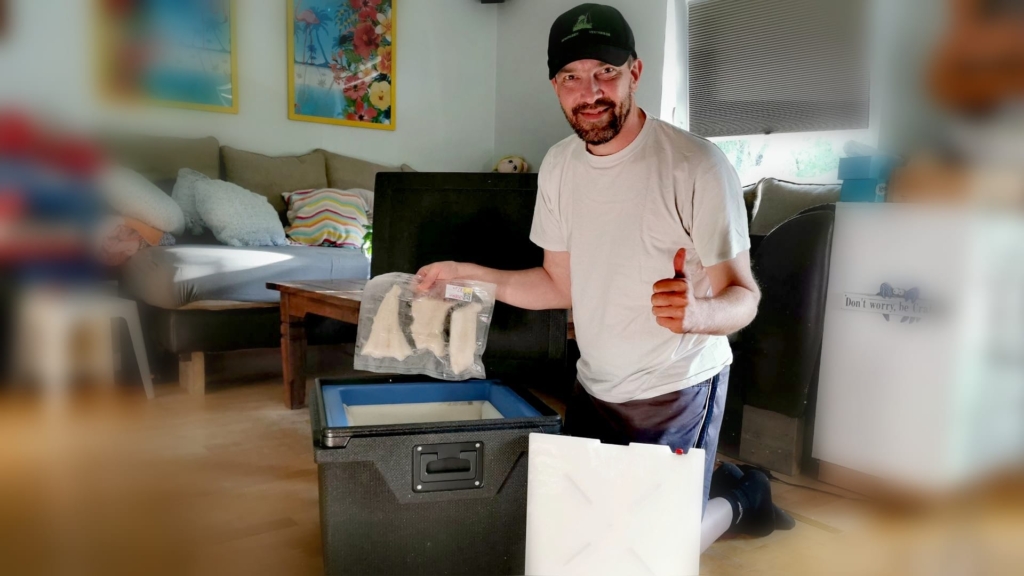 Ein Angler hält ein Paket mit tiefgefrorenen Fischfilets in der Hand, die er gerade aus einer QOOL-Box genommen hat - mit der anderen Hand signalisiert er durch einen Daumen nach oben, dass der Fisch nach vielen Tagen immer noch tief gefroren ist