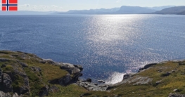 Uferspot zum Angeln am Akkarfjord - Blick vom Parkplatz auf das offene Meer, Sonnenglitzern auf der kräuselnden See und begrünte Felsen am Ufer
