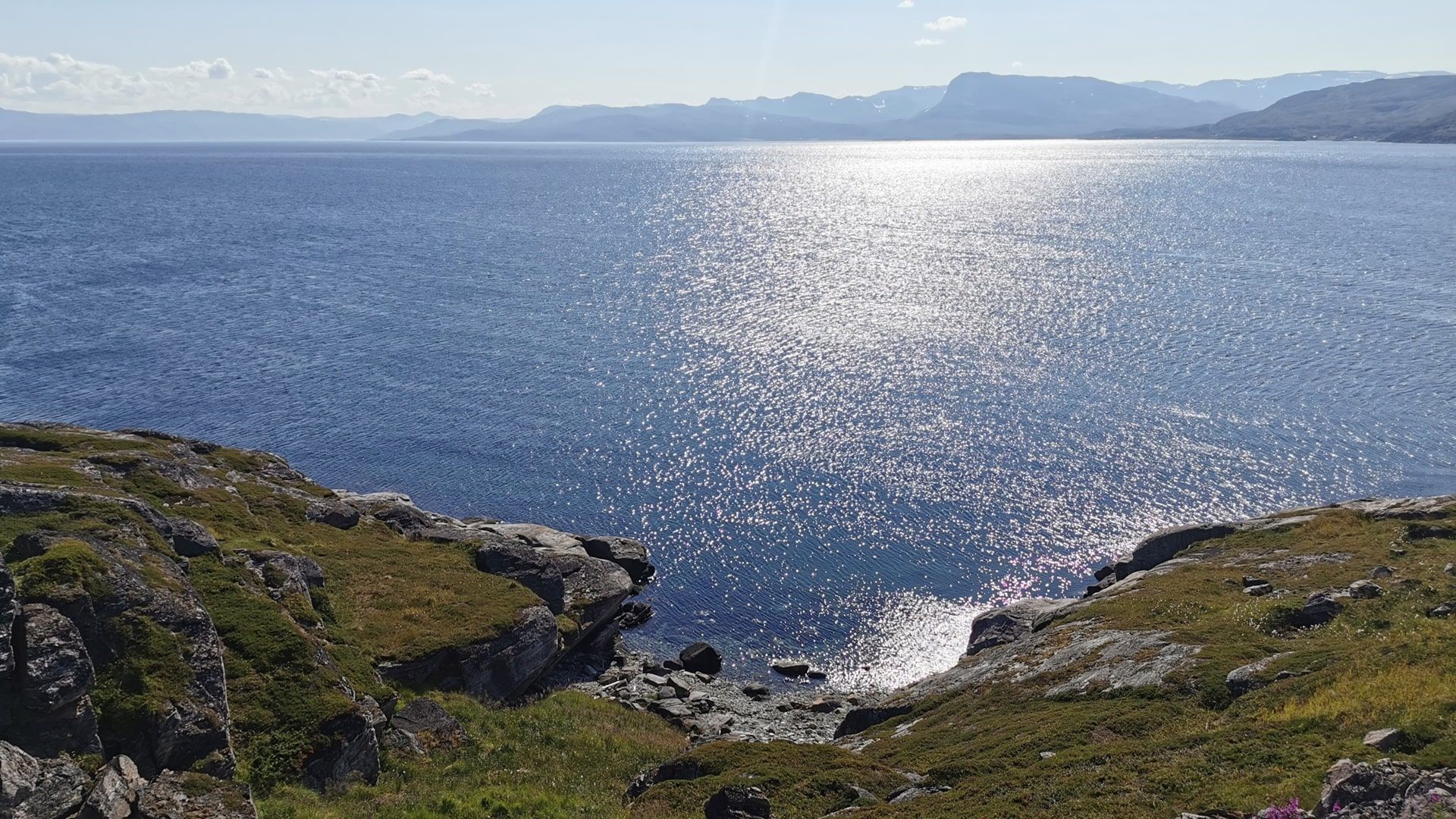 Uferspot zum Angeln am Akkarfjord - Blick vom Parkplatz auf das offene Meer, Sonnenglitzern auf der kräuselnden See und begrünte Felsen am Ufer