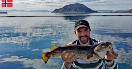 Ein freudiger Angler präsentiert seinen Dorsch, gefangen beim Angeln in Rypefjord – im Hintergrund spiegeln sich Insel und bewölkter Himmel in der glatten See