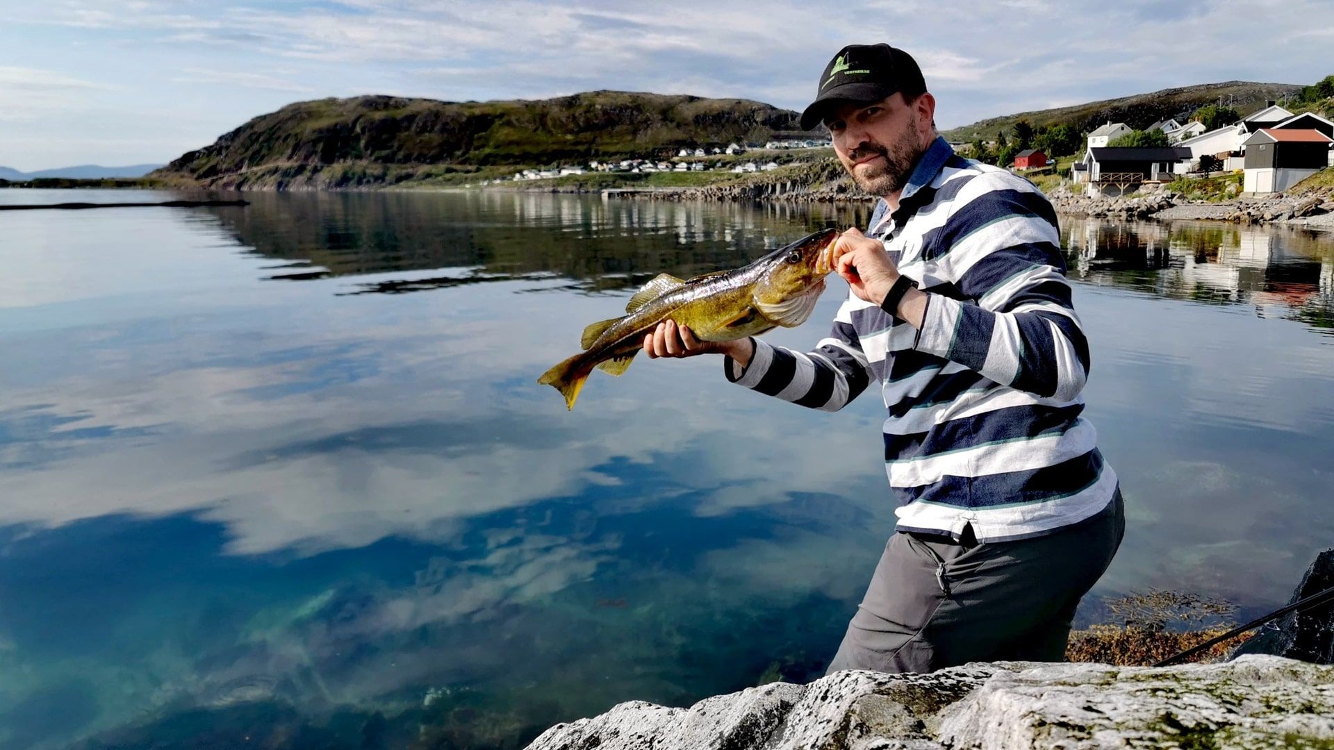 Angler mit Dorsch beim Angeln in Rypefjord – im Hintergrund das Ufer mit Häusern und ruhiger See unter leicht bewölktem Himmel
