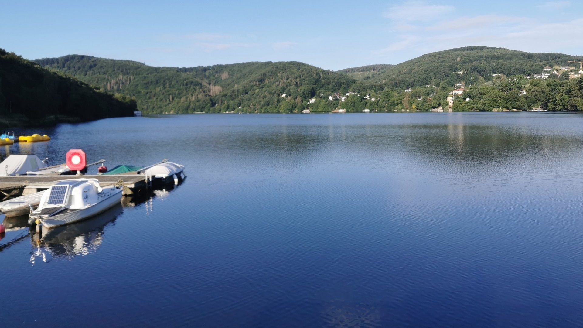 Blick von der Staumauer über das Staubecken der Rur bei Obermaubach an einem klaren Tag mit blauem Himmel – links der kleine Anleger des Bootsverleihs, im Hintergrund die sanften Hügel der Voreifel; ein beliebter Ort zum Angeln