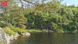 Blick entlang des felsigen, begrünten Ufers entlang der nördlichsten Insel im Øvstevatnet bei Lyngdal, an dem das Ufer direkt steil abfällt. Vor blauem Himmel ragt ein Kiefernzweig ins Bild.