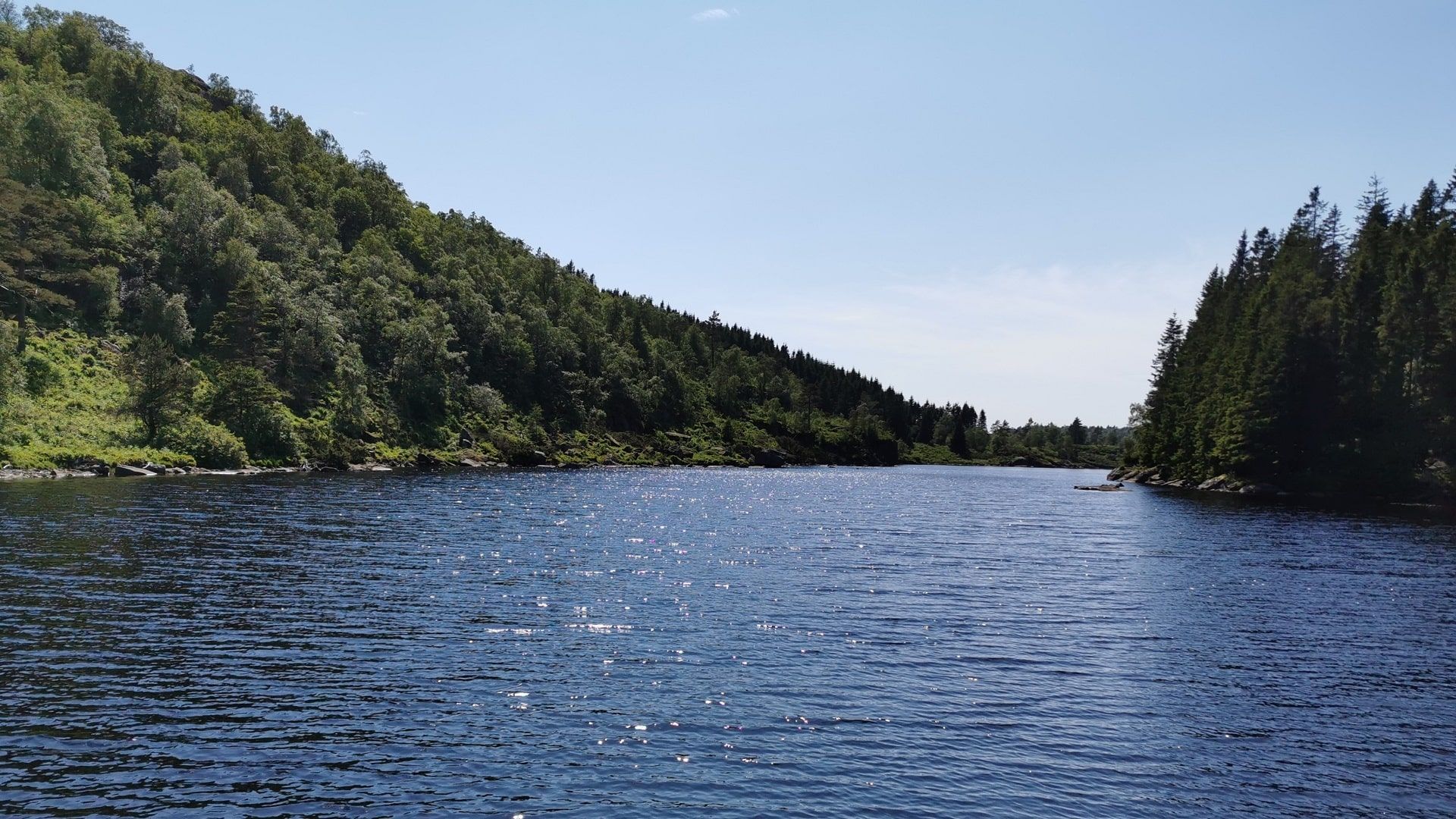 Panoramablick vom Nord- zum Südufer des Øvstevatnet. Sonnenlicht spiegelt sich im funkelnden Wasser mit leichten Wellen, umgeben von grünen Hängen.