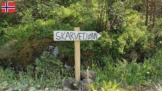 Ein Holzschild mit der Aufschrift "Skarvetjønn" zeigt den Weg nach rechts, umgeben von saftig grün bewachsenen Felsen.