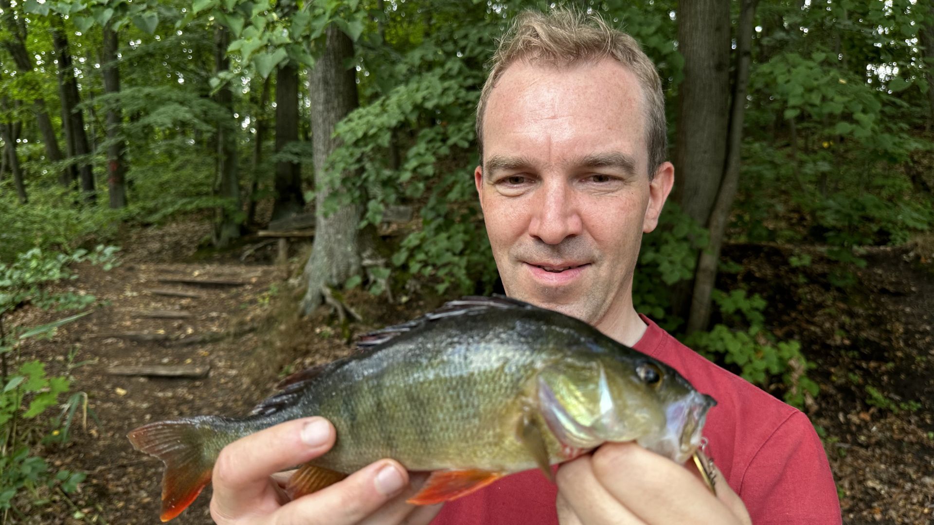 Ein Angler hält einen kleinen Barsch in die Kamera – gefangen mit einem goldenen Mepps-Spinner beim Spinnfischen am Trebuser See bei Fürstenwalde.