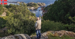 Von einem Felsen nahe Nesvåg hat ein Angler einen guten Blick und zeigt genau auf den Spot zum Angeln - die Hafeneinfahrt, die sich wie ein Minifjord mit begrünten Ufern unter blauem Himmel in den kleinen Ort erstreckt