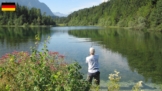 Angler am naturbelassenen Waldstausee der oberen Isar bei Krün, spiegelglattes, kristallklares Wasser, Nadelwaldinsel in der Ferne, Alpenpanorama und blühende Uferpflanzen bei strahlend blauem Himmel laden zum Angeln ein