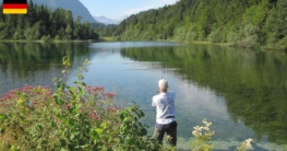 Angler am naturbelassenen Waldstausee der oberen Isar bei Krün, spiegelglattes, kristallklares Wasser, Nadelwaldinsel in der Ferne, Alpenpanorama und blühende Uferpflanzen bei strahlend blauem Himmel laden zum Angeln ein