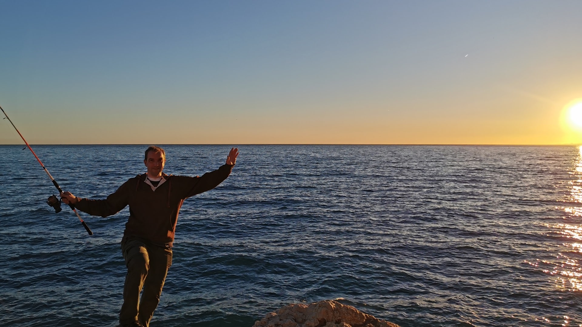 Ein Angler beim Spinnfischen im Osten von Málaga, hinter ihm blauer Himmel, vom Sonnenuntergang orange-rot gesäumt