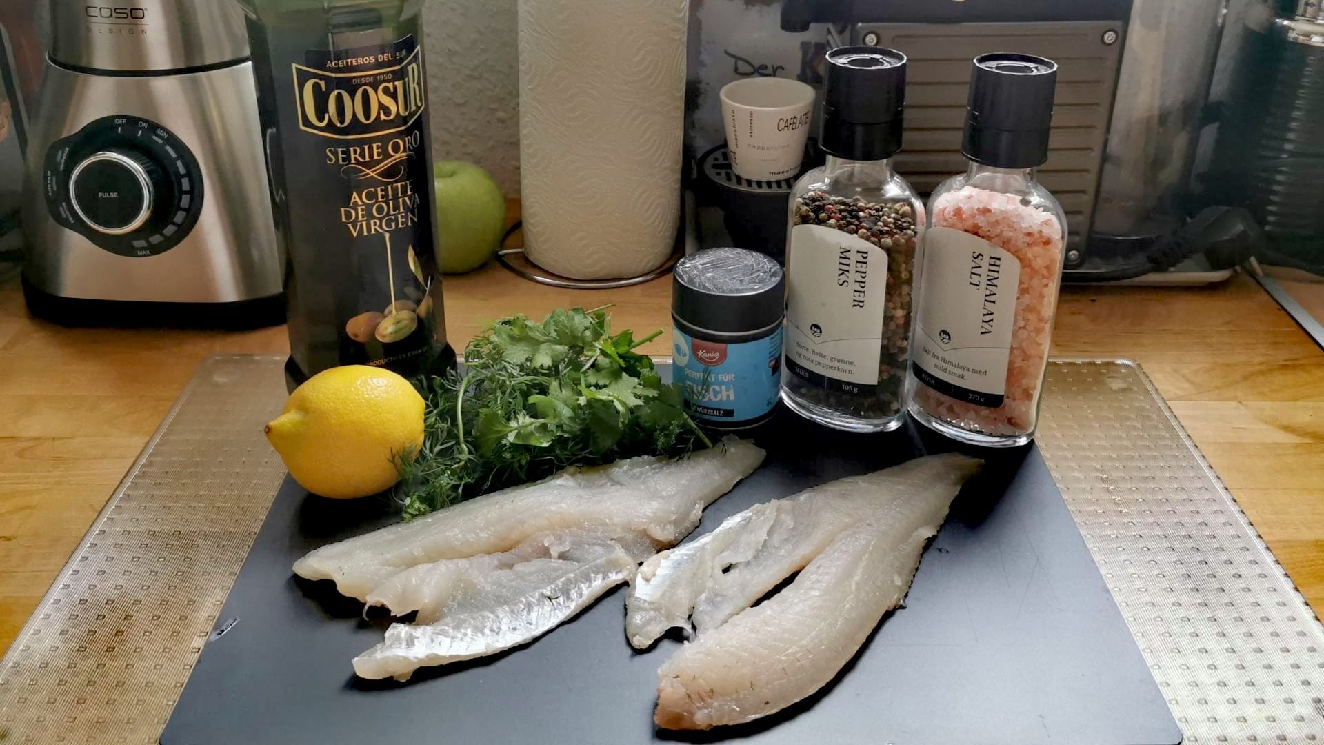 Zutaten für das Barschfilet aus dem Airfryer: Zwei große Barschfilets mit Gewürzen, Zitrone, Olivenöl, Dill und Koriander auf einem Schneidebrett angerichtet