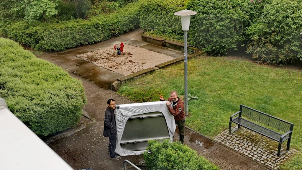 Zwei Personen tragen das in Laken eingepackte Dachzelt aus dem Keller – draußen beginnt die eigentliche Dachzelt-Montage.