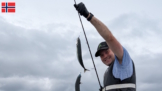 Ein Angler hält stolz seine Rute mit einem Makrelen-Paternoster in die Kamera – gleich mehrere Makrelen hängen daran; im Hintergrund zieht sich ein dichter, bewölkter Himmel über die norwegische Küste.