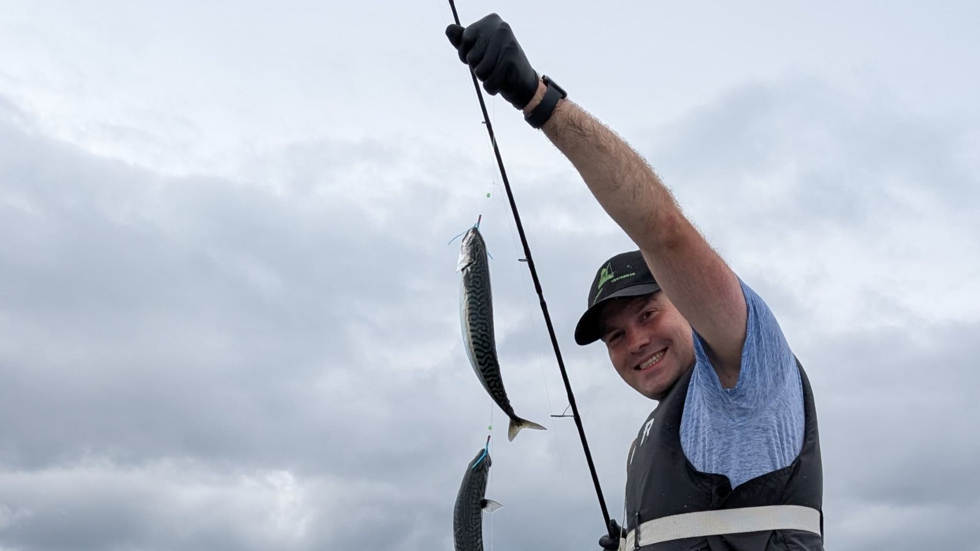 Ein Angler hält stolz seine Rute mit einem Makrelen-Paternoster in die Kamera – gleich mehrere Makrelen hängen daran; im Hintergrund zieht sich ein dichter, bewölkter Himmel über die norwegische Küste.