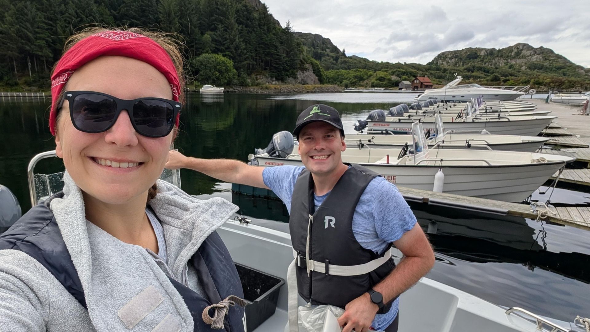 Zwei gut gelaunte Angler kurz vor dem Auslaufen mit einem Leihboot in Hausvikstranda bei Korshamn. Die Vorfreude auf den Offshore-Trip ist spürbar – rundherum weitere Boote, grüner Hang im Hintergrund, spiegelglattes Wasser im Hafen.