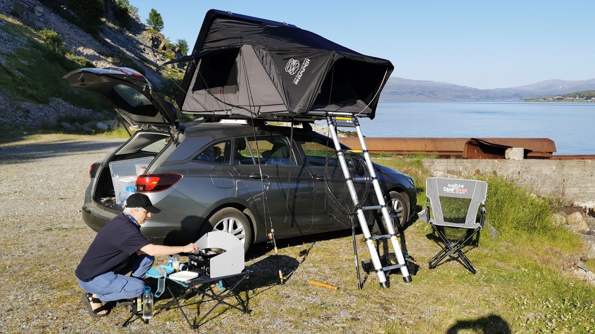 Reise-Alltag am Kvalsund: Vor dem geöffneten Dachzelt bereitet eine Frau die mobile Campingküche vor
