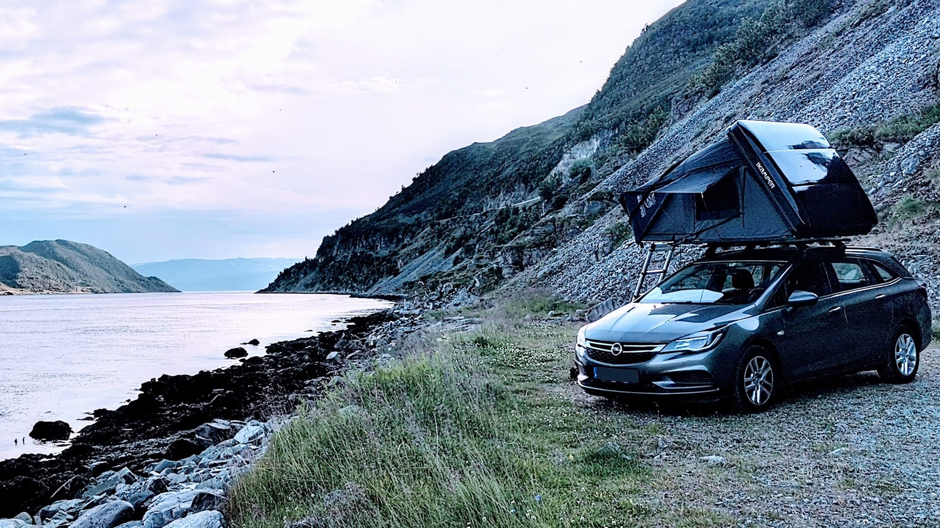 Auto mit Dachzelt auf einem Felsplateau unter der Kvalsundbrua, ausgerichtet zum offenen Meer nach Westen