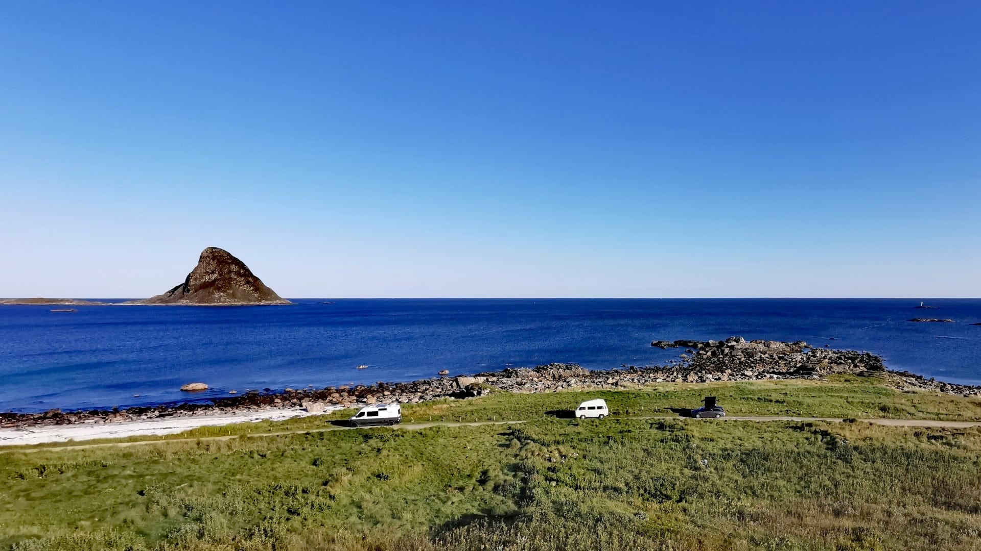 Panoramablick auf den Stellplatz am Bleik Sandstrand: weißer Sand, grüne Wiese, tiefblaues Meer und eine haiflossenförmige Felsinsel