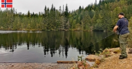 Angler beim Angeln oberhalb der Staumauer des Flekkerøvann bei Skien, spiegelglattes Wasser, rötlicher Felsboden und von Mischwald gesäumtes Ufer