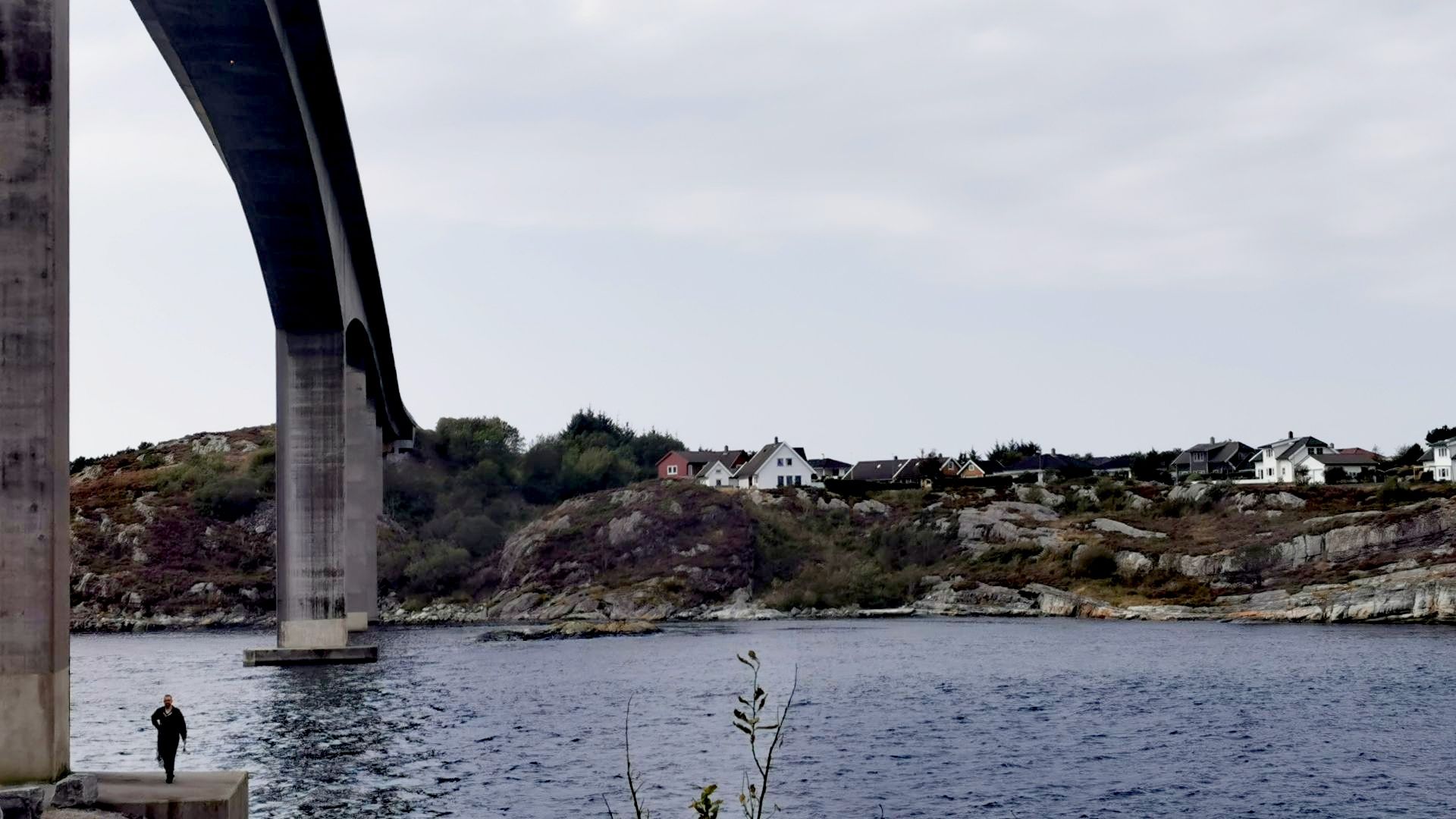 Angler beim Angeln am Rongesundet auf Sotra bei Bergen, winzig klein auf dem Sockel der imposanten Brücke, gegenüber typische norwegische Häuser am Ufer