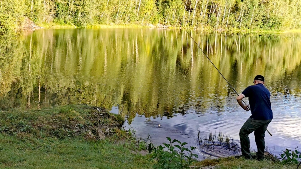 Autor drillt einen Fisch mit der Savage Gear Parabellum CCS 10'1 Test-Rute an einem sonnigen, windstillen norwegischen Bergsee, grüne Ufer spiegeln sich im Wasser.