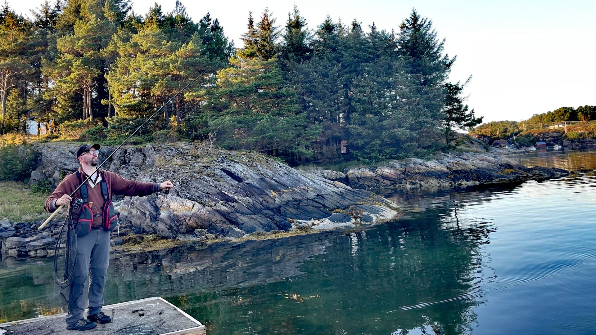 Autor hält die Savage Gear Parabellum CCS 10'1 Test-Rute in der rechten Hand und zieht mit der linken an der Schnur, um die semiparabolische Biegekurve zu prüfen, Hintergrund ruhige Felsküste mit Nadelbäumen.