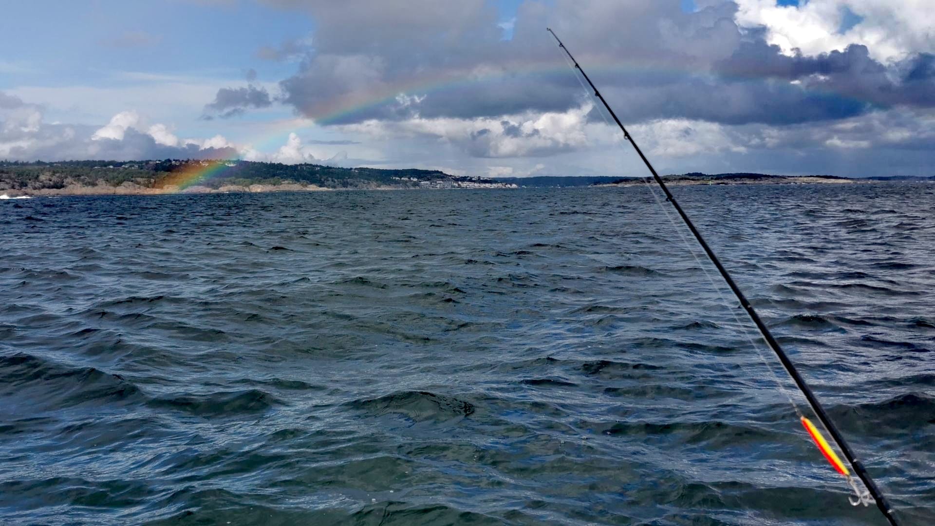 Blick vom Boot aufs Wasser bei Risør mit Wellen, Regenbogen und grüner Küste; im Vordergrund eine Angelrute mit rot-orangener Pilker