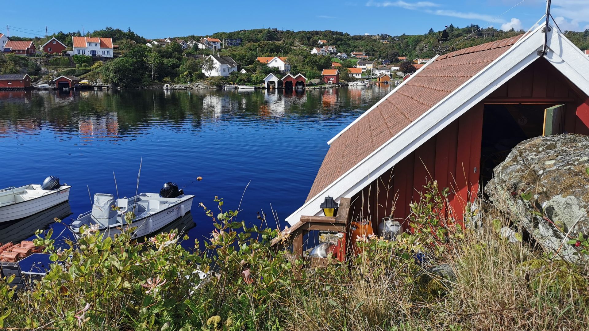 Rotes Bootshaus direkt am Wasser auf der Insel Skjernøya bei Mandal, mit blauem Himmel und mehreren festgemachten Booten.