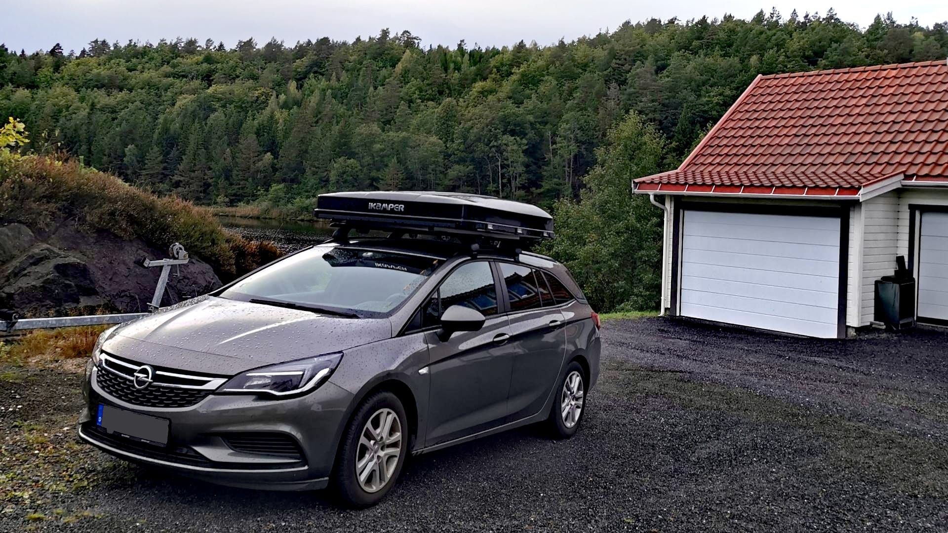 Auto mit zugeklapptem Dachzelt vor der Unterkunft in Risør bei der Abfahrt nach Mandal; feuchtes Wetter, geschotterte Fläche und Nadelwald im Hintergrund.