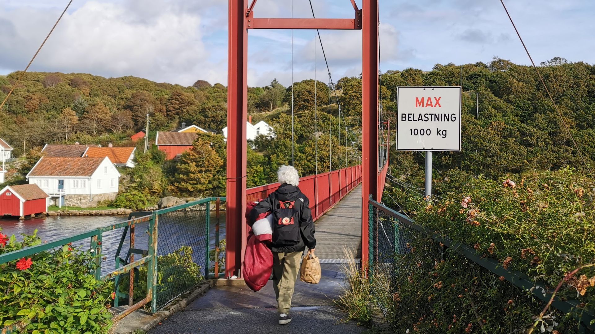 Frau mit Gepäck betritt die rote Hängebrücke auf Skjernøya; ein Schild weist die maximale Belastung von 1000 Kilogramm aus.