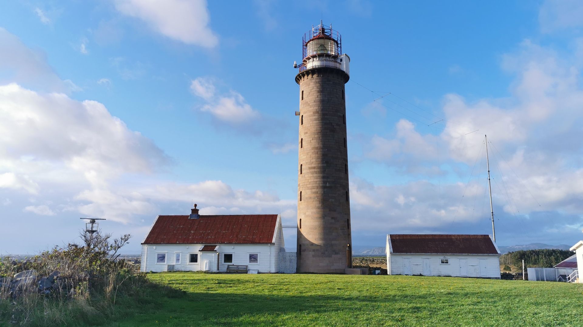 Listafyr nahe Lyngdal, ein weißer Leuchtturm mit grünem Rasen und kleinen Häusern unter blauem Himmel mit Wolken