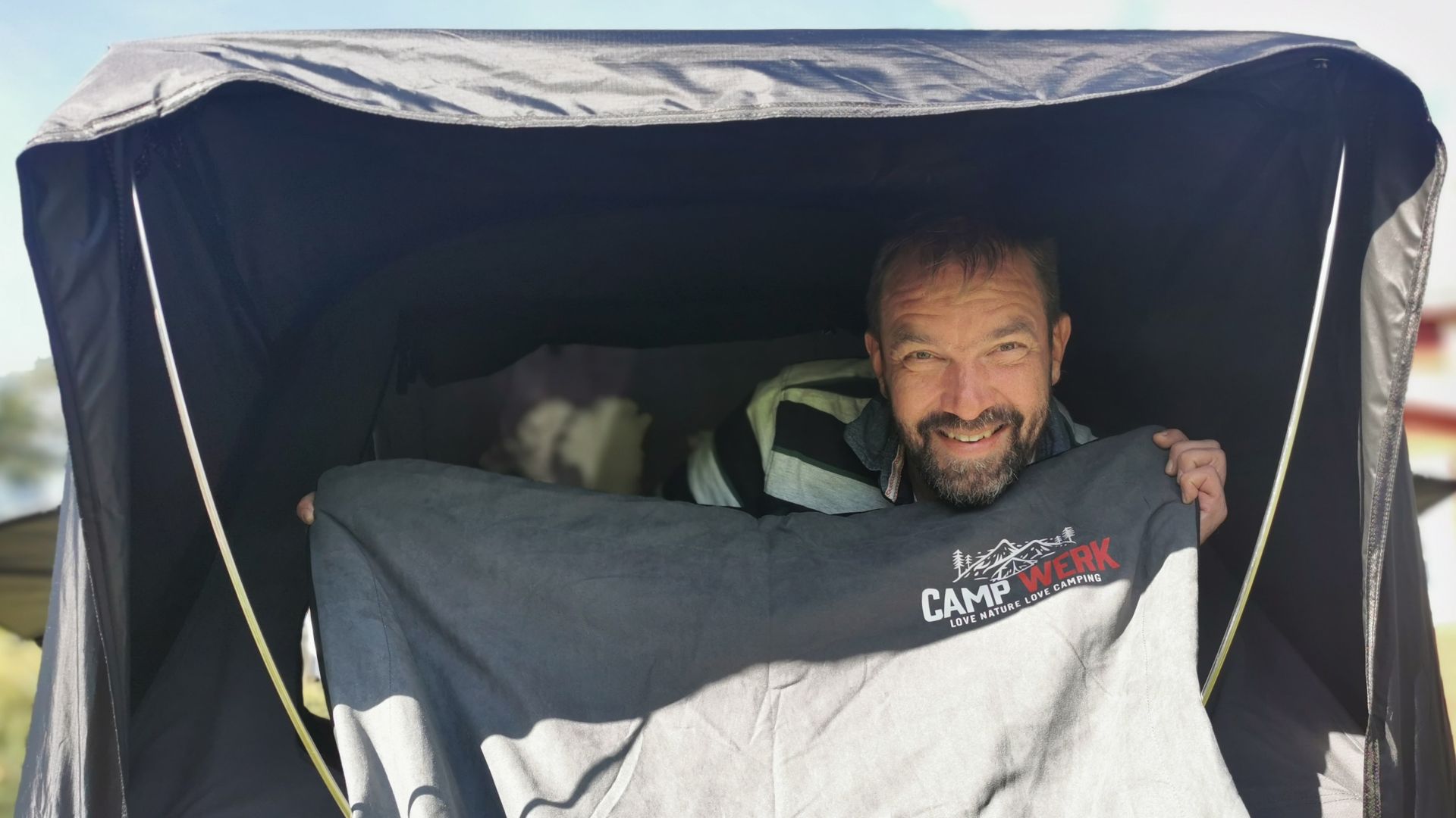 Autor hält lächelnd eine Campwerk-Heizdecke aus dem geöffneten Dachzelt, betrieben mit Powerbank für Wärme in frostiger Nacht